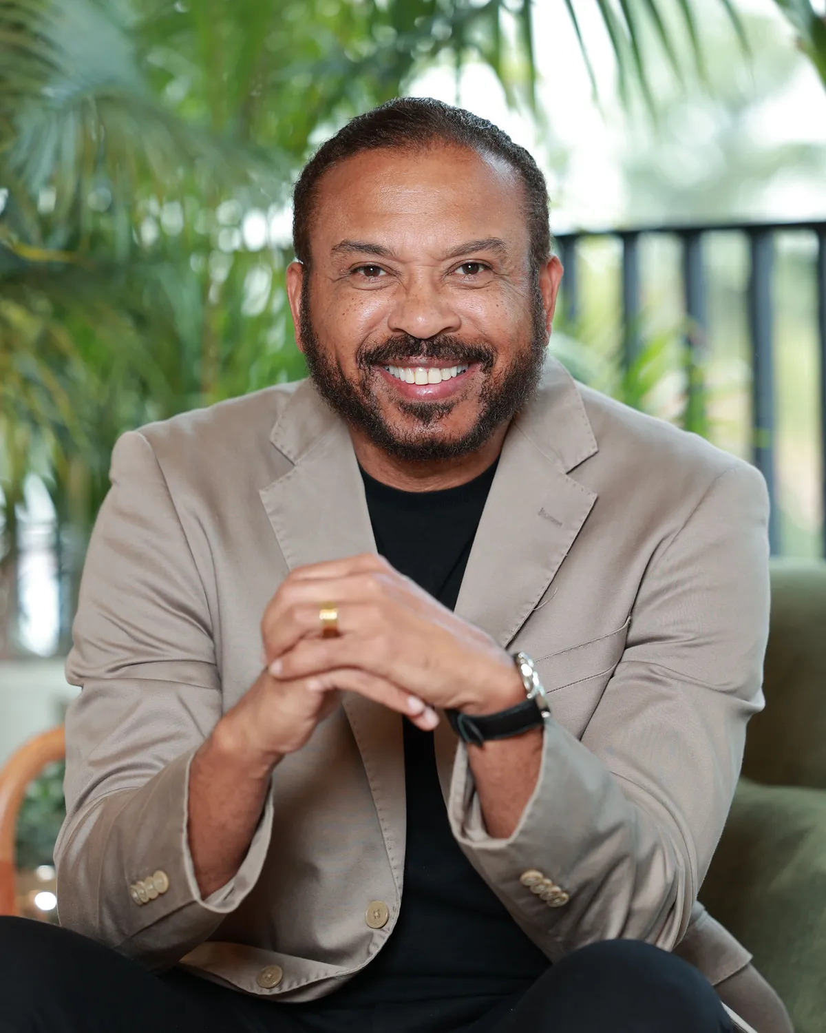 Dr. Ribamar Cruz sorrindo, de blazer bege, sentado em um ambiente com plantas.